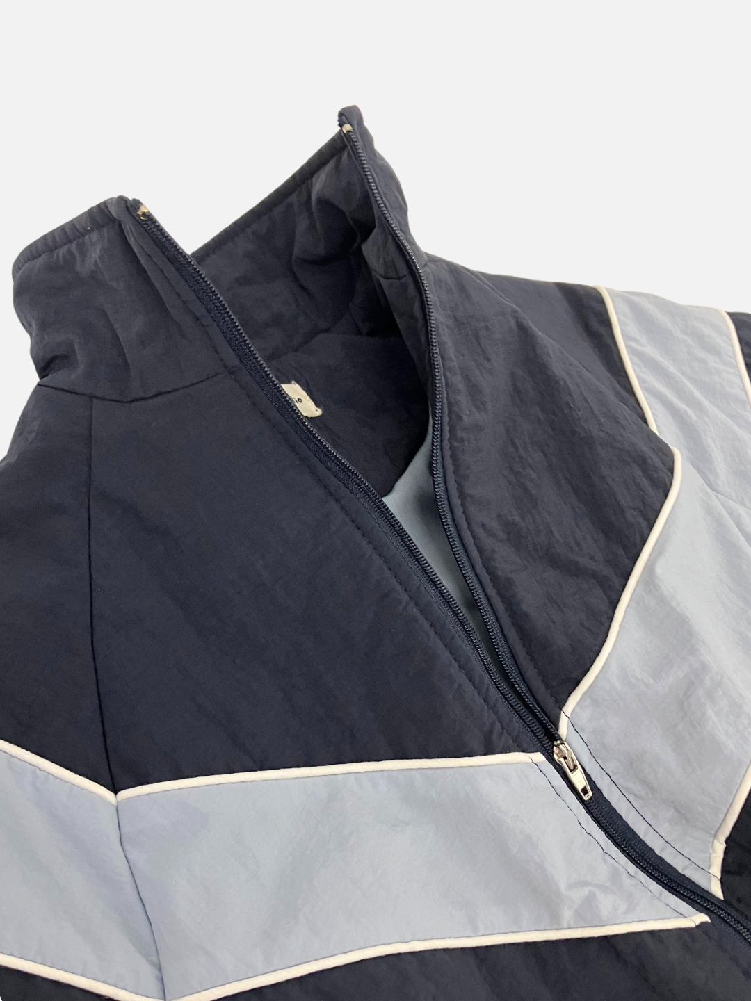 Navy | Close-up of the WARM-UP JACKET, a navy and light grey windbreaker, reminiscent of a retro track jacket, featuring a partially open front zipper and a small Korean label inside the collar.