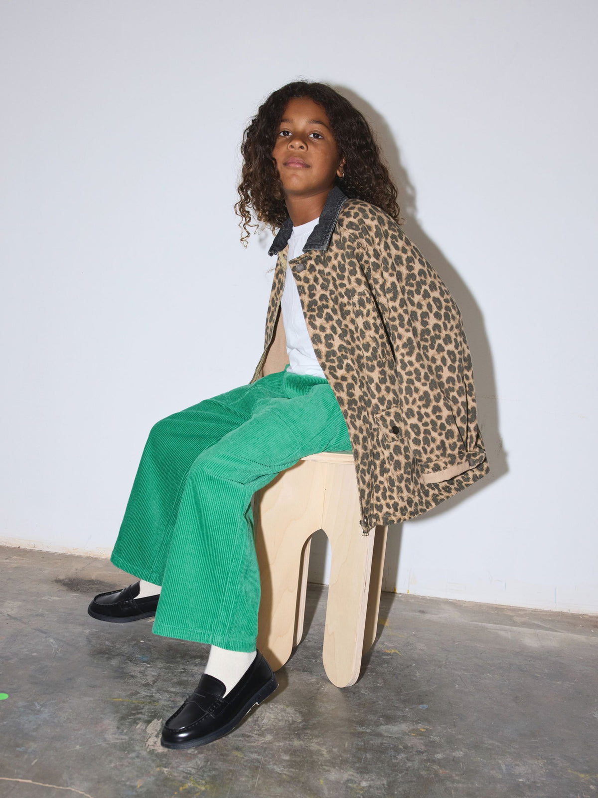 Leopard | A child sits on a light wood stool wearing a leopard print jacket, bright green corduroy trousers, a white t-shirt, and black loafers against a plain white wall.