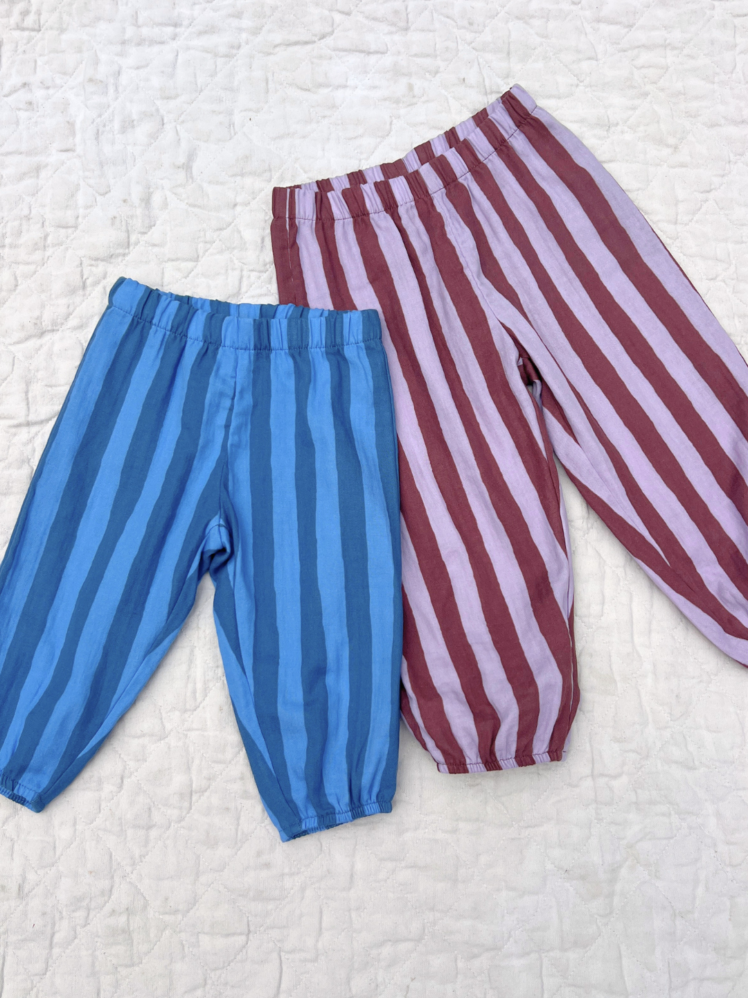 Two pairs of the baby and toddler organic muslin pants, one blue striped and one purple and maroon striped, laid flat on a beige quilt.