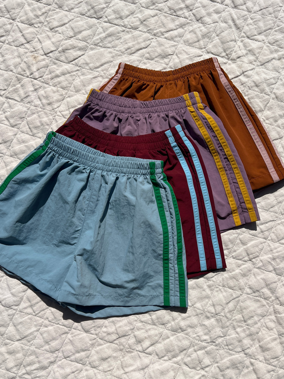 Blue | Four pairs of SPRINTER SHORTS in blue, maroon, lavender, and brown with contrasting side stripes and elasticated waistbands are displayed overlapping on a quilted white surface.