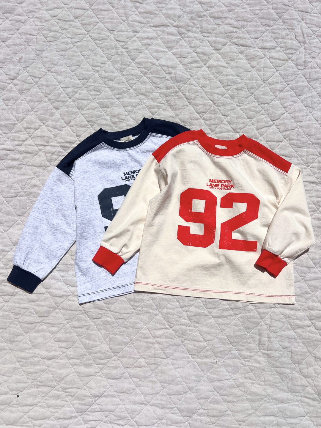 Navy/Light Heather | Two MEMORY LANE LONGSLEEVE tees from a Korean kids brand lay flat on a quilted surface: one light gray with navy accents, the other cream with red accents and a vintage sports jersey “92” printed on the front.