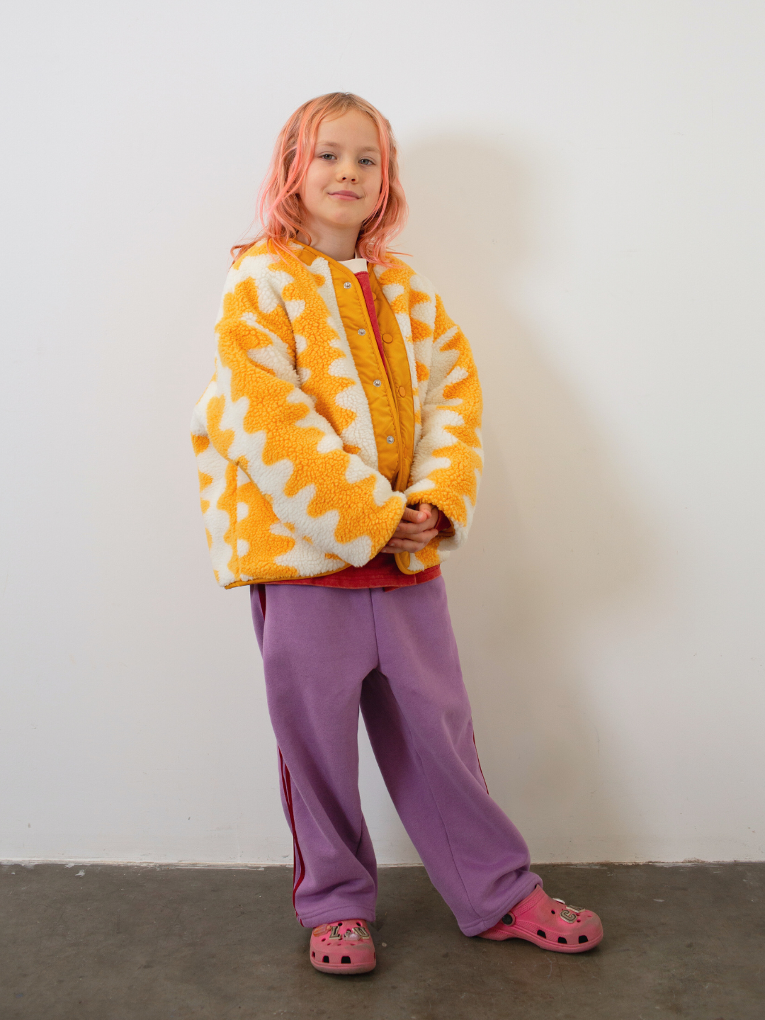 Child wearing a yellow and white patterned jacket with purple pants against a plain background