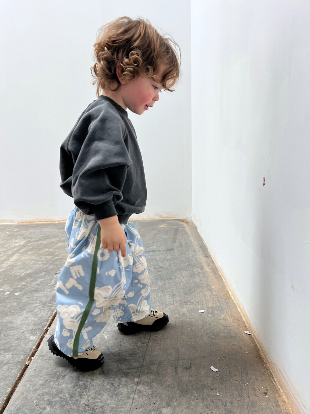 A young child with curly hair, wearing a dark sweatshirt, SKYWRITING PANT, and black shoes, stands on a concrete floor near a white wall.