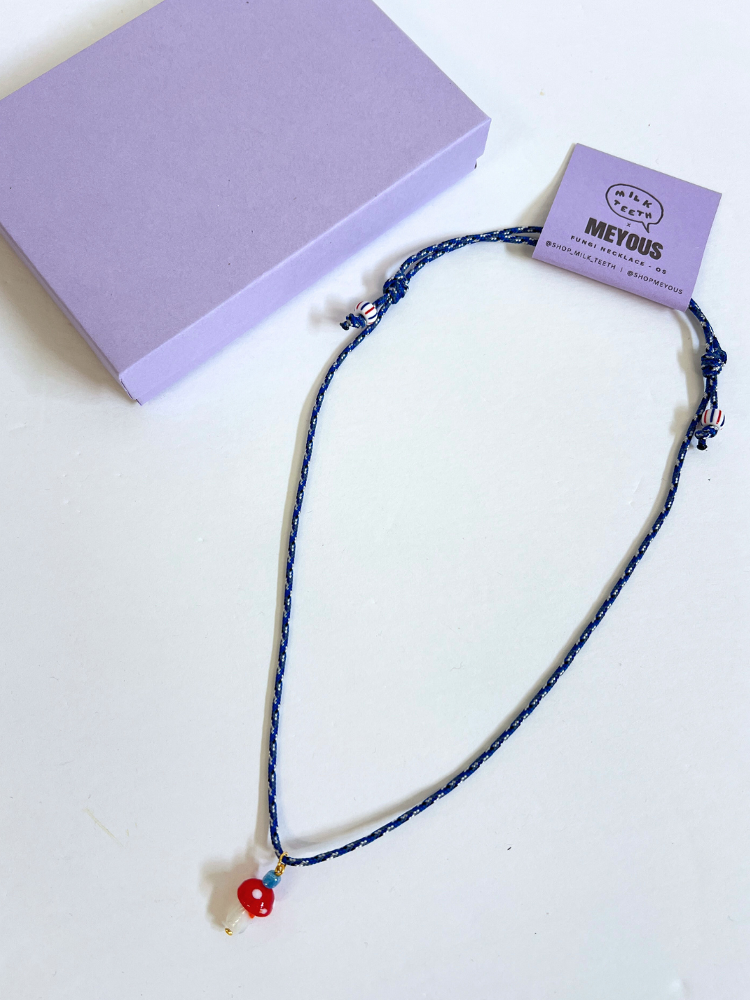 The FUNGI NECKLACE, a blue braided piece with a glass mushroom charm pendant, is displayed on a white surface beside a closed lavender box, highlighting the playful elegance of handmade genderless jewelry from Meyous jewelry.