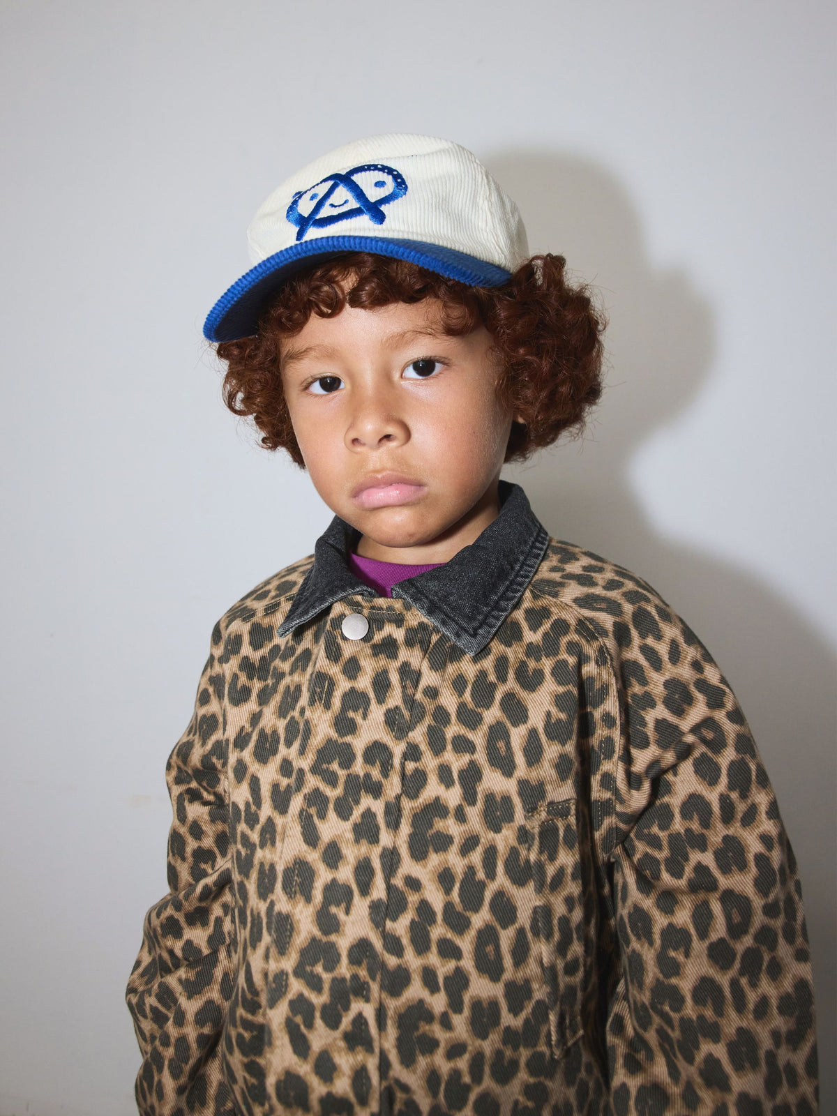 Leopard | A child wearing a leopard-print shirt jacket with a dark denim collar, a white and blue baseball cap, and curly brown hair, photographed against a plain light gray wall.
