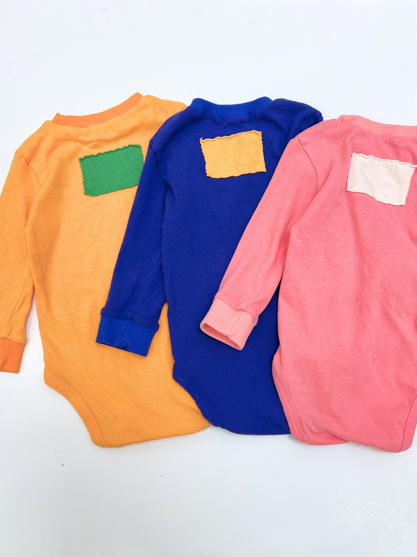 PATCH ONESIE: Three long-sleeve onesies from a Korean brand in orange, blue, and pink, made of soft cotton and featuring a colorful patch on the upper back, are displayed flat on a white surface.