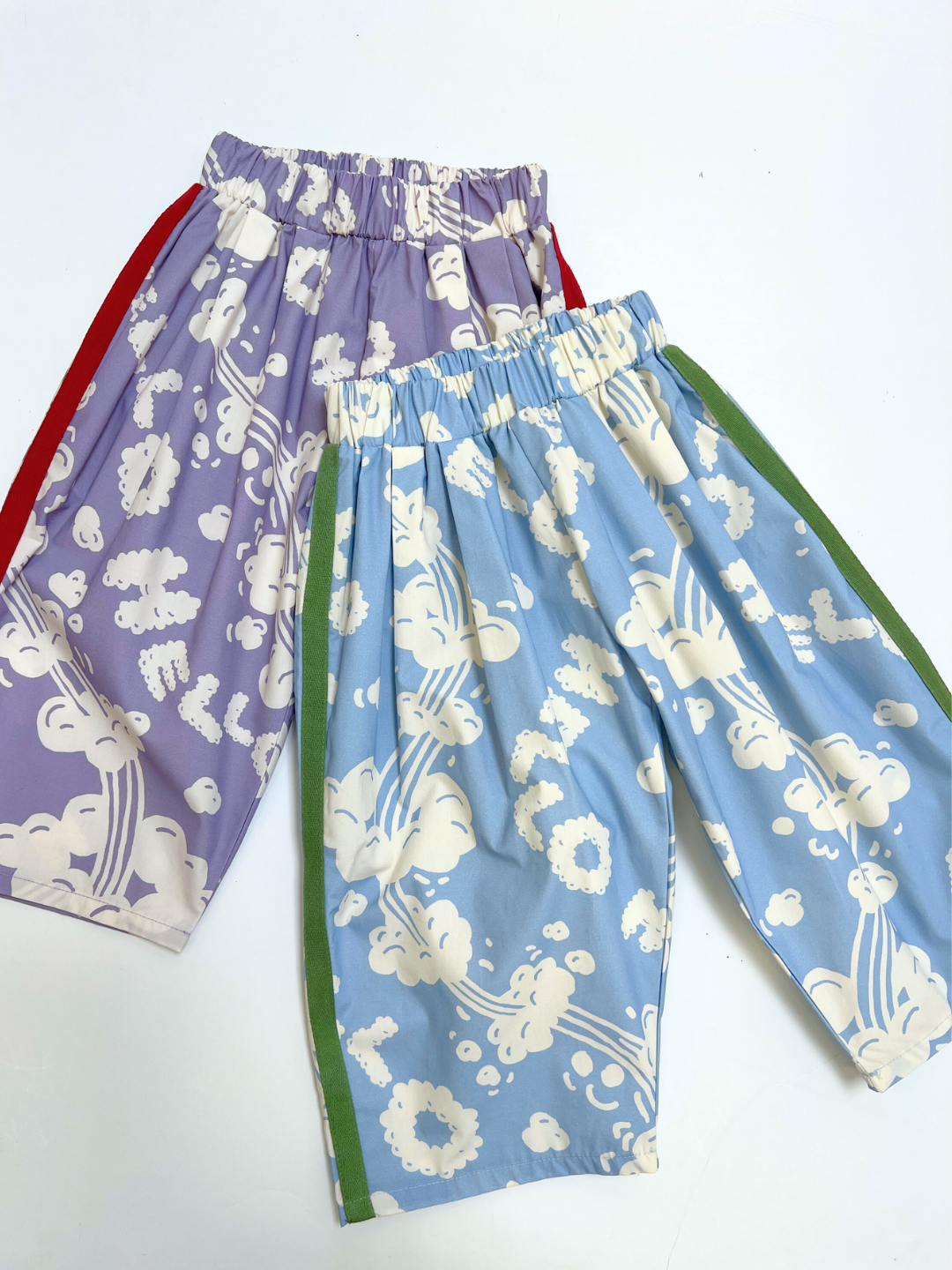 Bleu | Two pairs of SKYWRITING PANT elastic-waist shorts with cloud patterns—one lavender with red side stripes and one light blue with green stripes—are displayed on a white background.