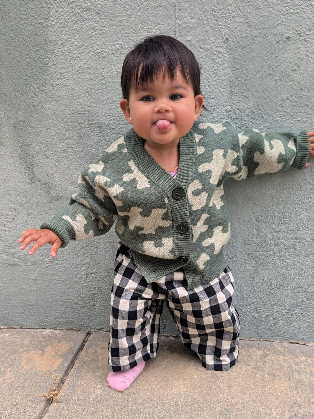 Green | A baby stands against a gray wall, wearing the PUZZLE CARDIGAN in green camouflage, black-and-white checkered pants, and a pink sock, playfully sticking out their tongue.