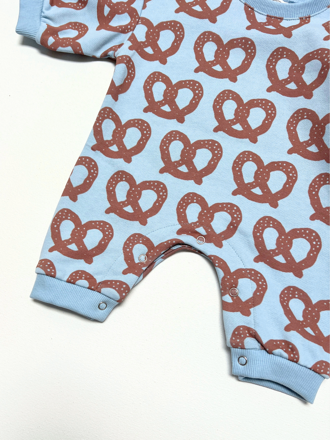 Brown/Blue | A baby romper in light blue with an all-over brown pretzel print, featuring ribbed cuffs and a snap-button crotch.