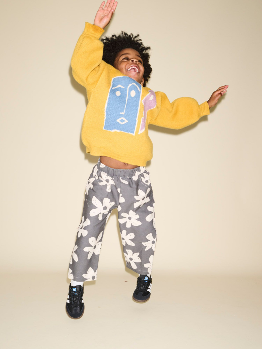 A child wearing the TEATIME SWEATER, featuring a whimsical face design, and patterned pants is caught mid-jump in front of a plain background, perfectly capturing the joy and spontaneity often sought in product photos.