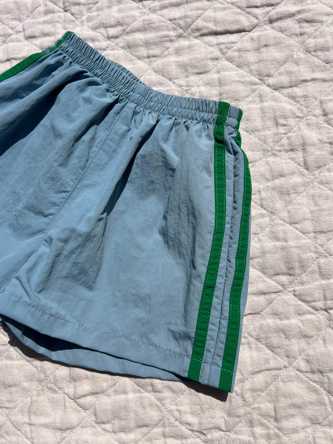 Blue | SPRINTER SHORTS in light blue with green side stripes and an elastic waist are displayed on a white quilted surface under natural light.