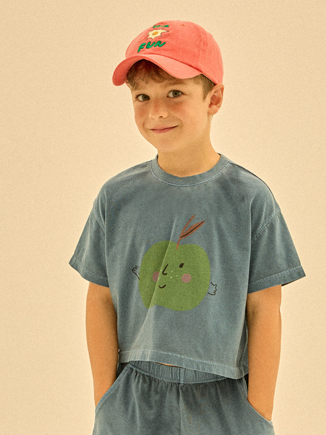 A young boy in a pink cap and blue outfit featuring an APPLE PIGMENT CROPPED T-SHIRT stands with his hands in his pockets, smiling.