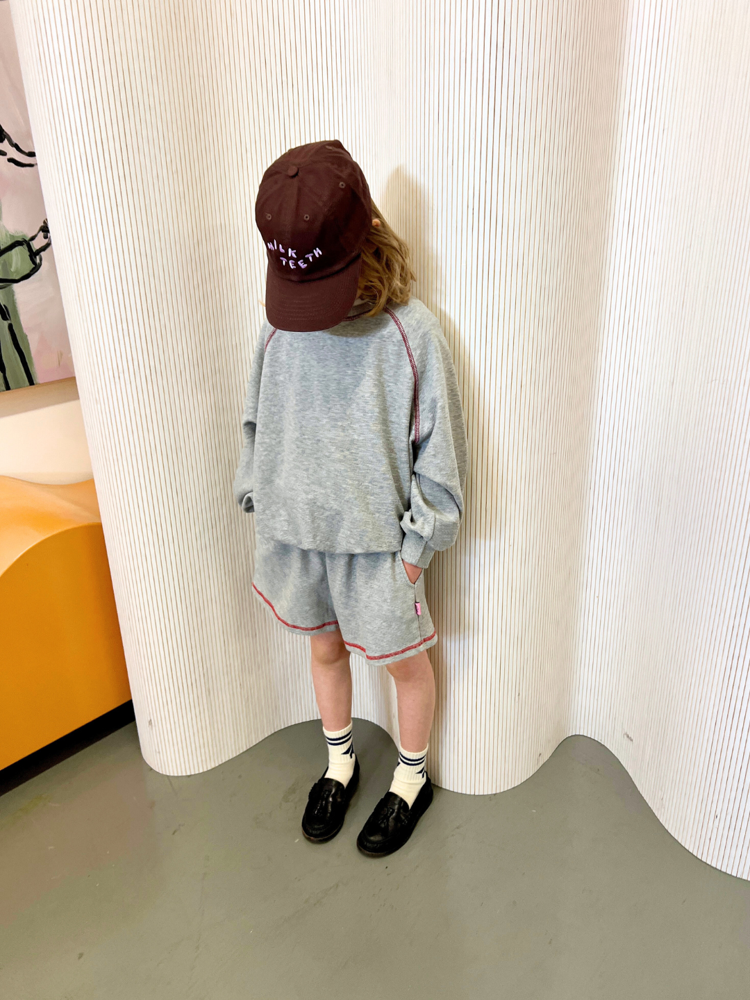 A child in a gray outfit and maroon cap looks down, standing against a light-striped curtain, wearing a retro-style VARSITY SWEATSHIRT by Cantucci Studios.