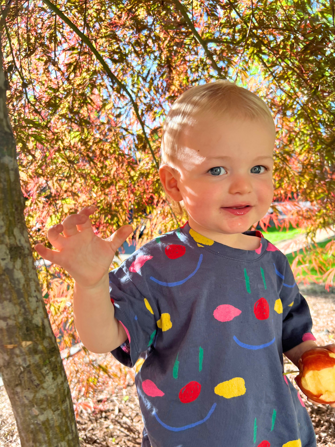 A toddler wearing the kids' Facepaint Tee in Navy, standing under a tree and holding an apple.