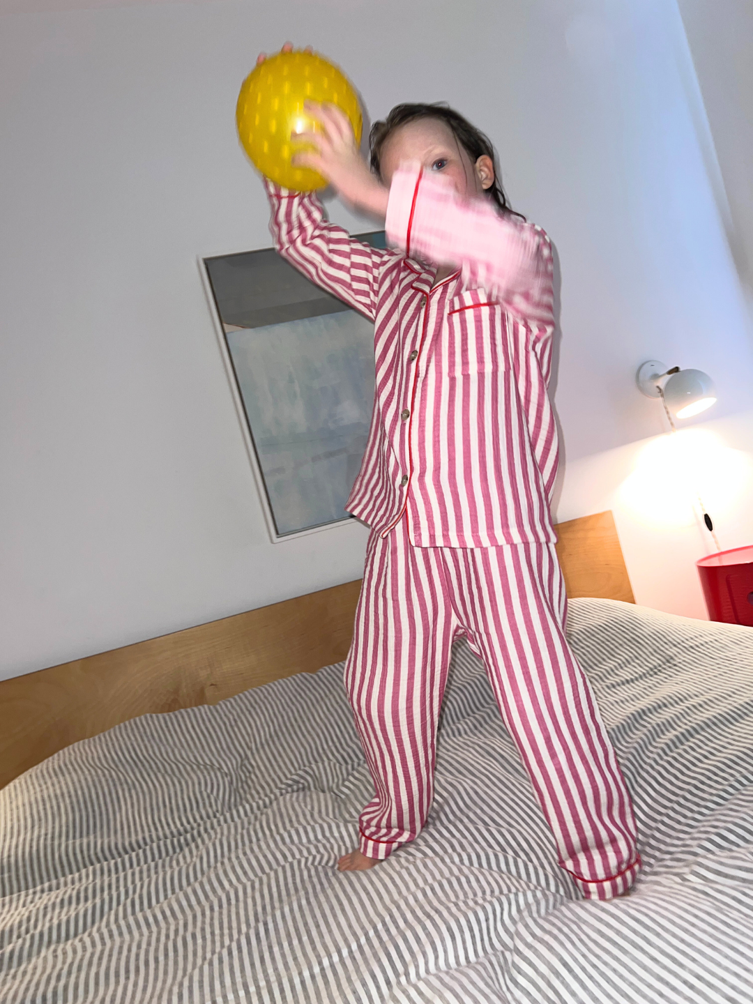 Pink Stripe | A child in the MOVIE NIGHT SET, featuring striped red and white set made from breathable fabric, stands on a bed holding a yellow ball. The cozy attire matches the warm ambiance with a painting and wall lamp in the background.