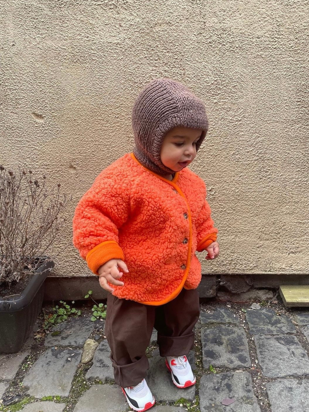 Tangerine | A toddler wearing an orange collarless fleece jacket with four brown buttons, with a brown hat, brown pants and white sneakers, standing on a paved outdoor floor, against a beige wall..