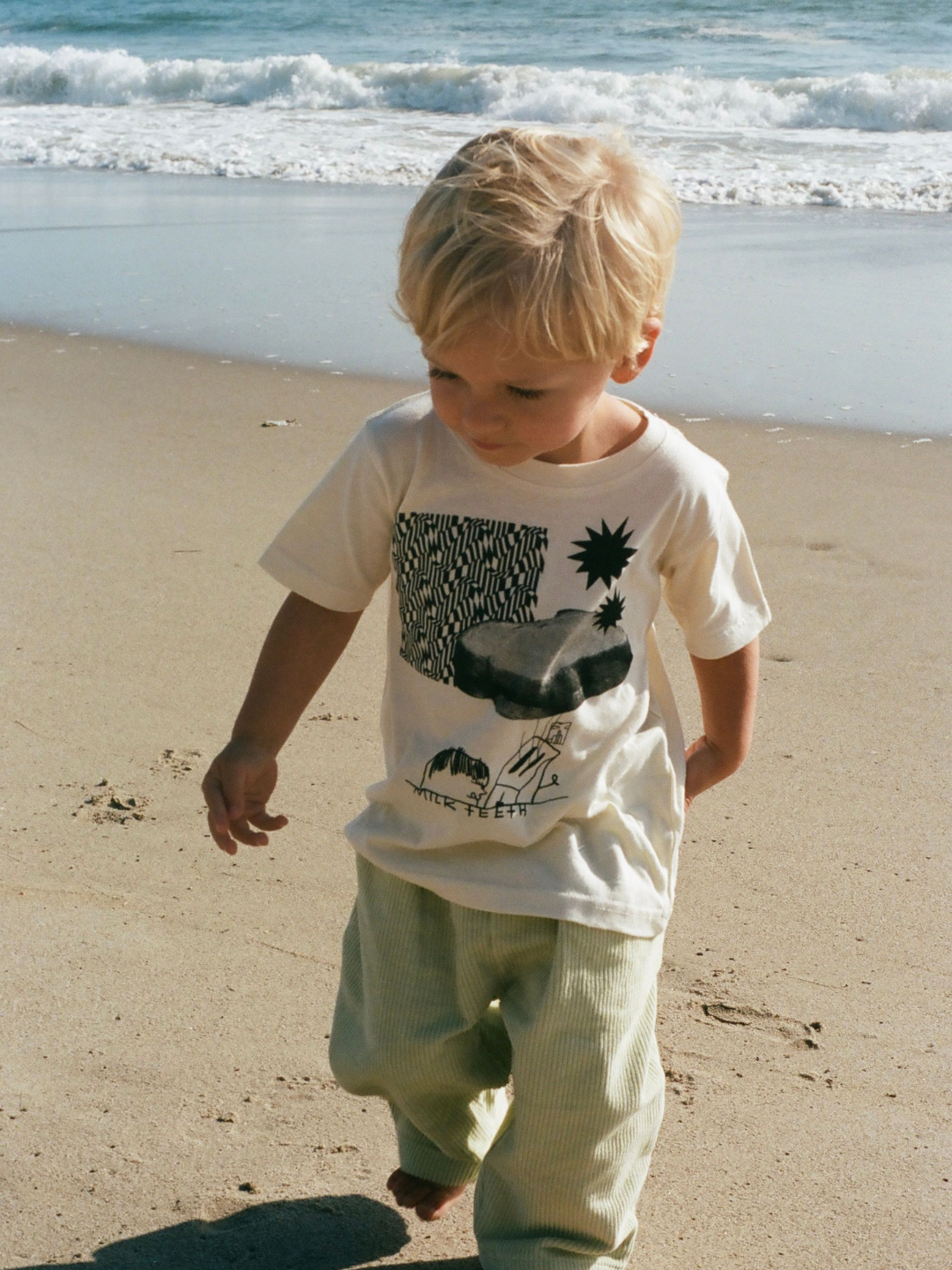 A child on a beach wearing a kids' tee shirt in cream, printed in black with stars, zigzags, a PBJ sandwich, a toaster and the words Milk Teeth