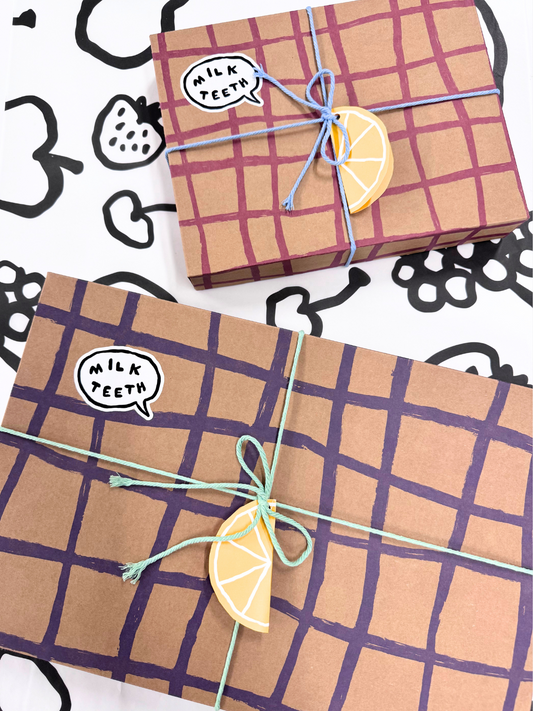 Image of Two GIFT WRAP boxes featuring a purple grid pattern, embellished with lemon slice decorations, blue string, and a "Milk Teeth" speech bubble sticker, arranged on a monochrome patterned surface. Inside each box is a charming gift card and specialty tissue paper for an extra touch of elegance.