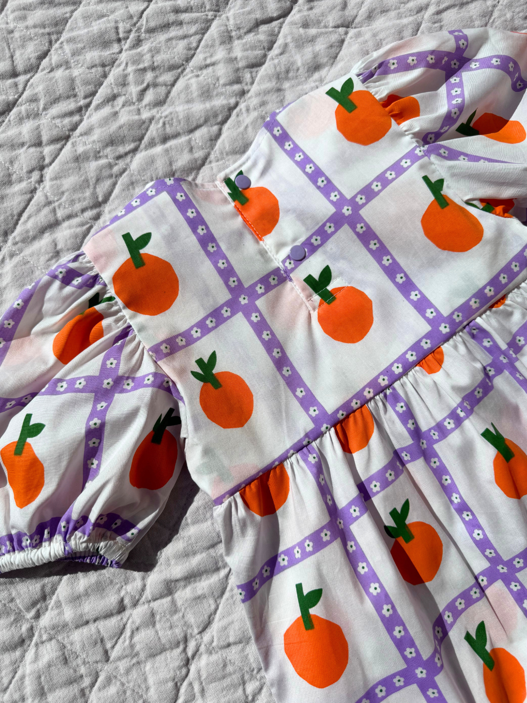 A close up of the back view of the kid's Glow dress with an all-over oranges print showcasing the snap closures