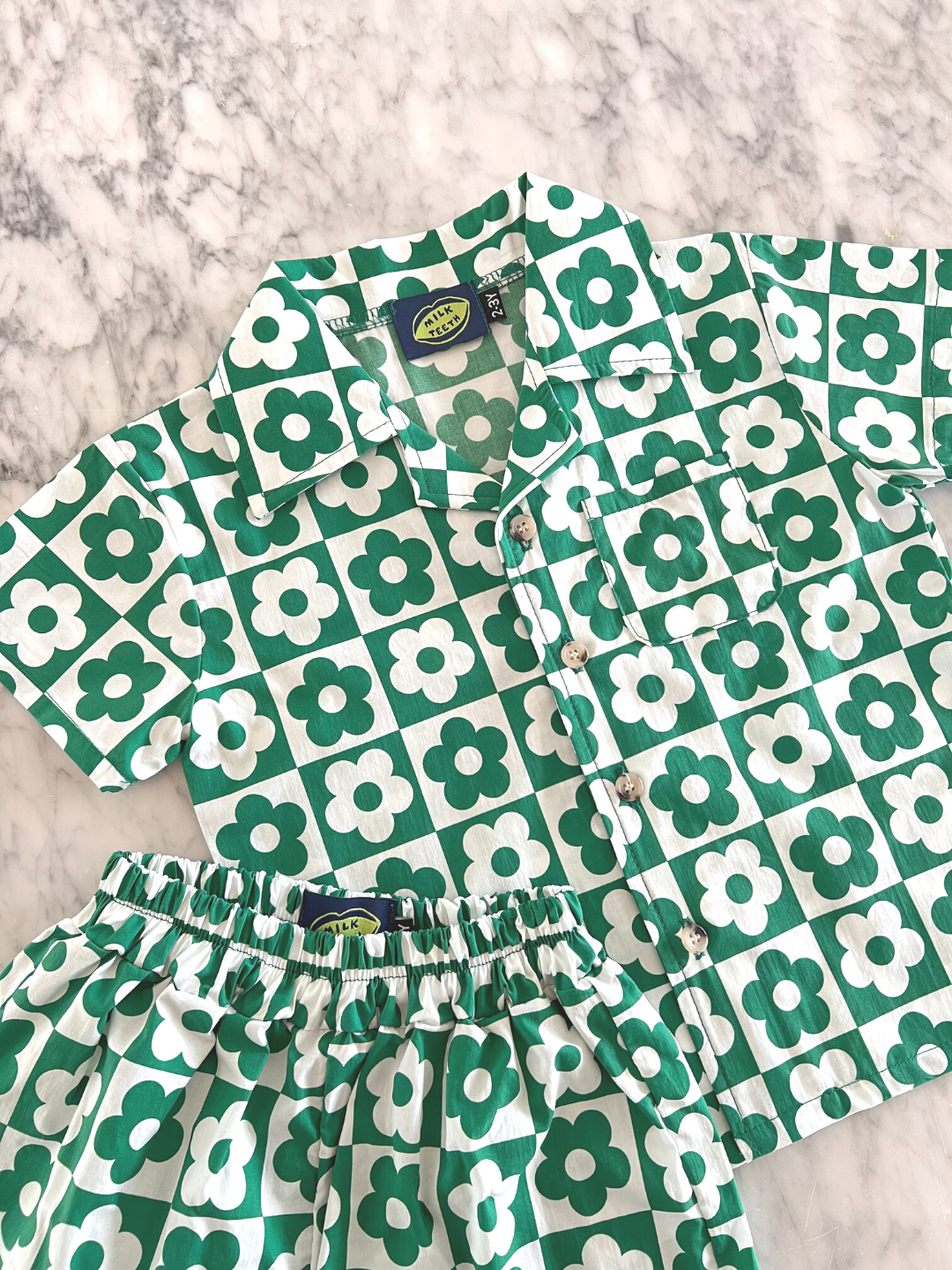 Pine | Kids' set of shirt and shorts in a checkerboard pattern of green and white flowers, laid on a table, front view