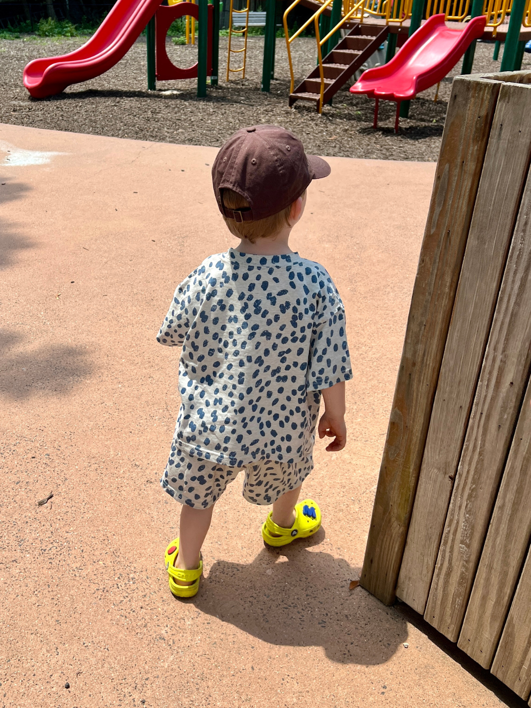 A toddler in a matching outfit featuring the "SPOTTED TEE" and yellow sandals, possibly from a Korean kids brand, stands near a large wooden structure in a playground. The relaxed t-shirt of the outfit adds comfort while the toddler faces away, looking toward the red slide and other playground equipment in the background.