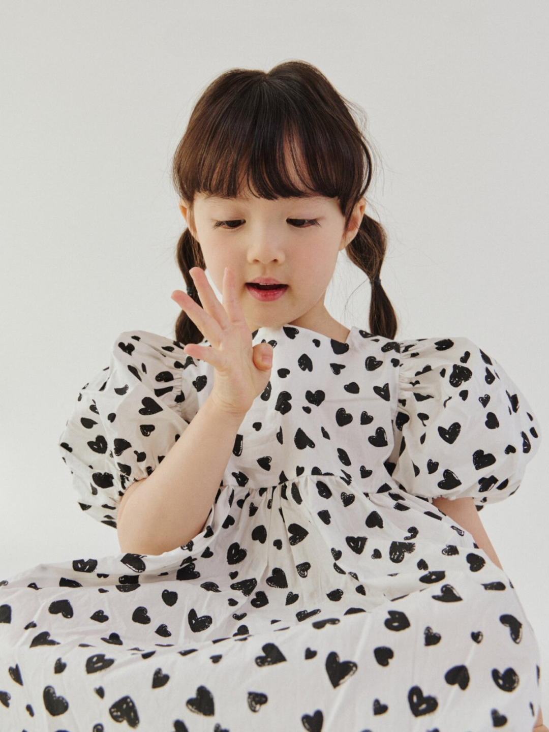 Seated child wearing a kids' puff-sleeved dress in a pattern of black hearts on a white background