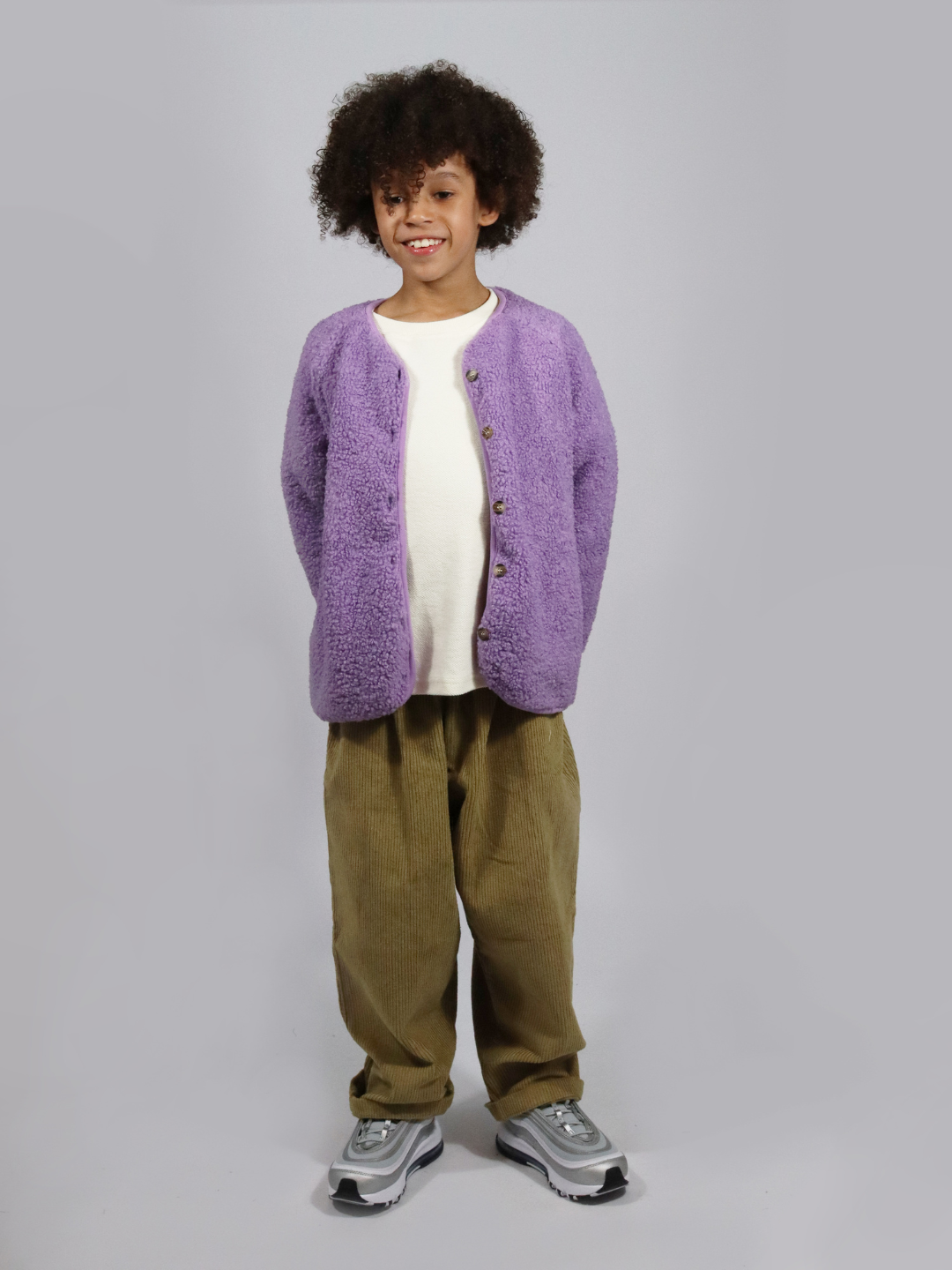 A child wearing a light purple collarless fleece jacket with four brown buttons, with olive green pants, a white t-shirt and grey sneakers, standing on a light grey background.