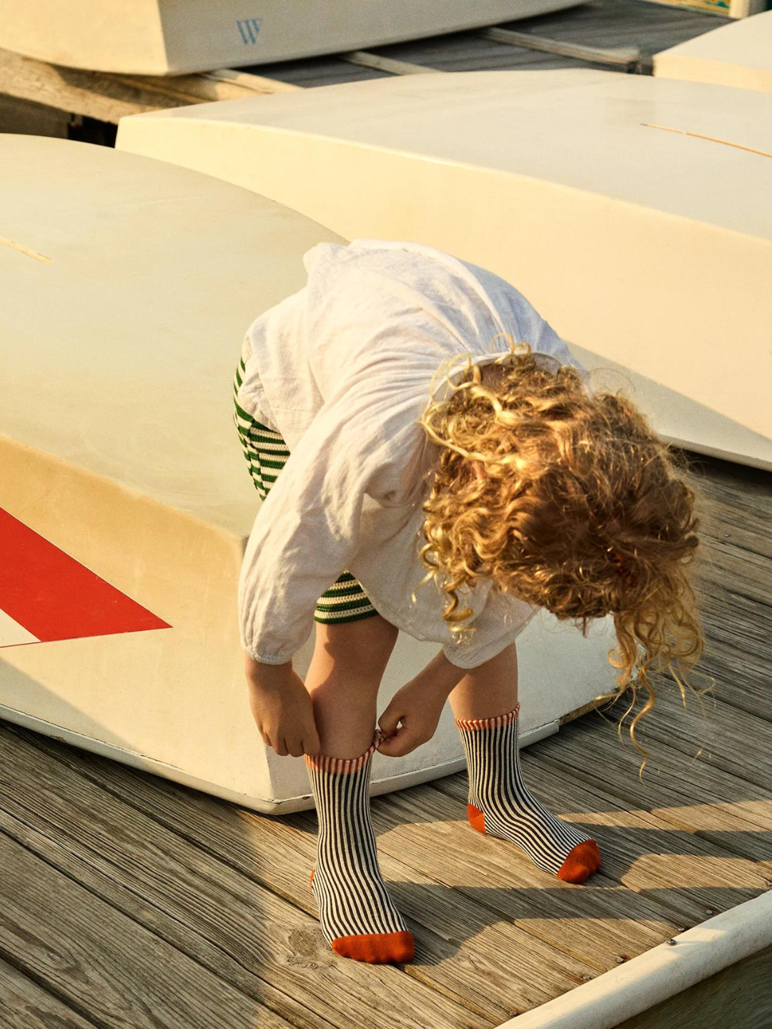 Black | A child wearing socks in narrow black and white vertical stripes with a solid red toe and heel, and red stripes at ankle cuff. Child is bending over near a boat, on a wooden deck, and wears a white shirt and green and cream striped shorts.