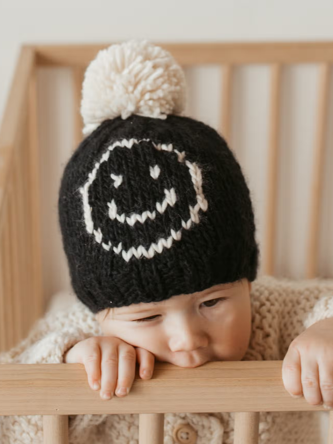 Baby wearing a black beanie hat in chunky yarn with an off-white smiley face on the front and an off-white pom-pom on top. The baby is show standing in a wooden crib, wearing a cream cardigan, and chewing the side of the crib.