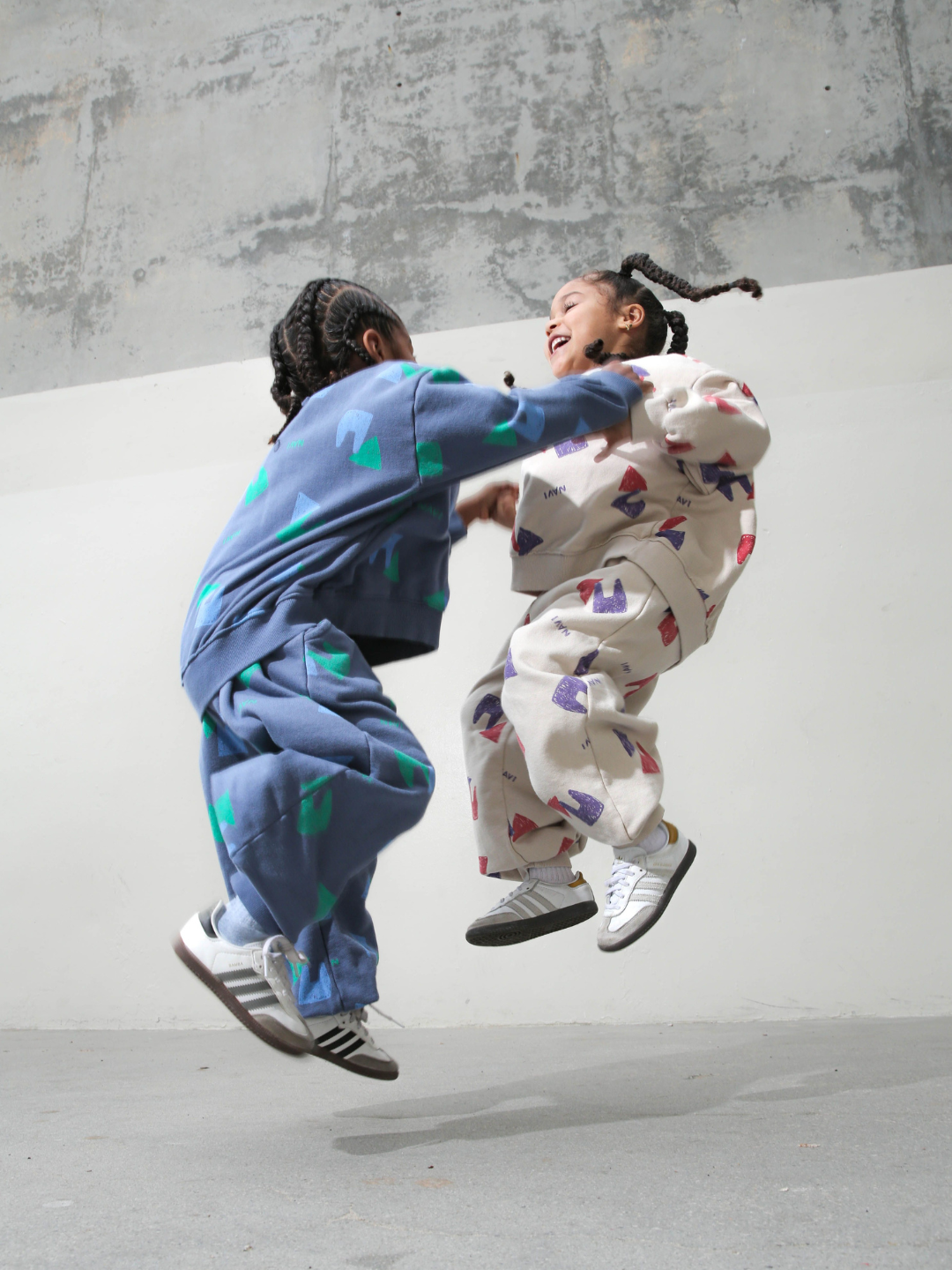 Child wearing blue sweatpants with an all over pattern of green and blue shapes and the brand name Navi, with matching crewneck sweatshirt. He is jumping in front of a grey background, holding hands with another child in the same outfit in the oatmeal colorway.