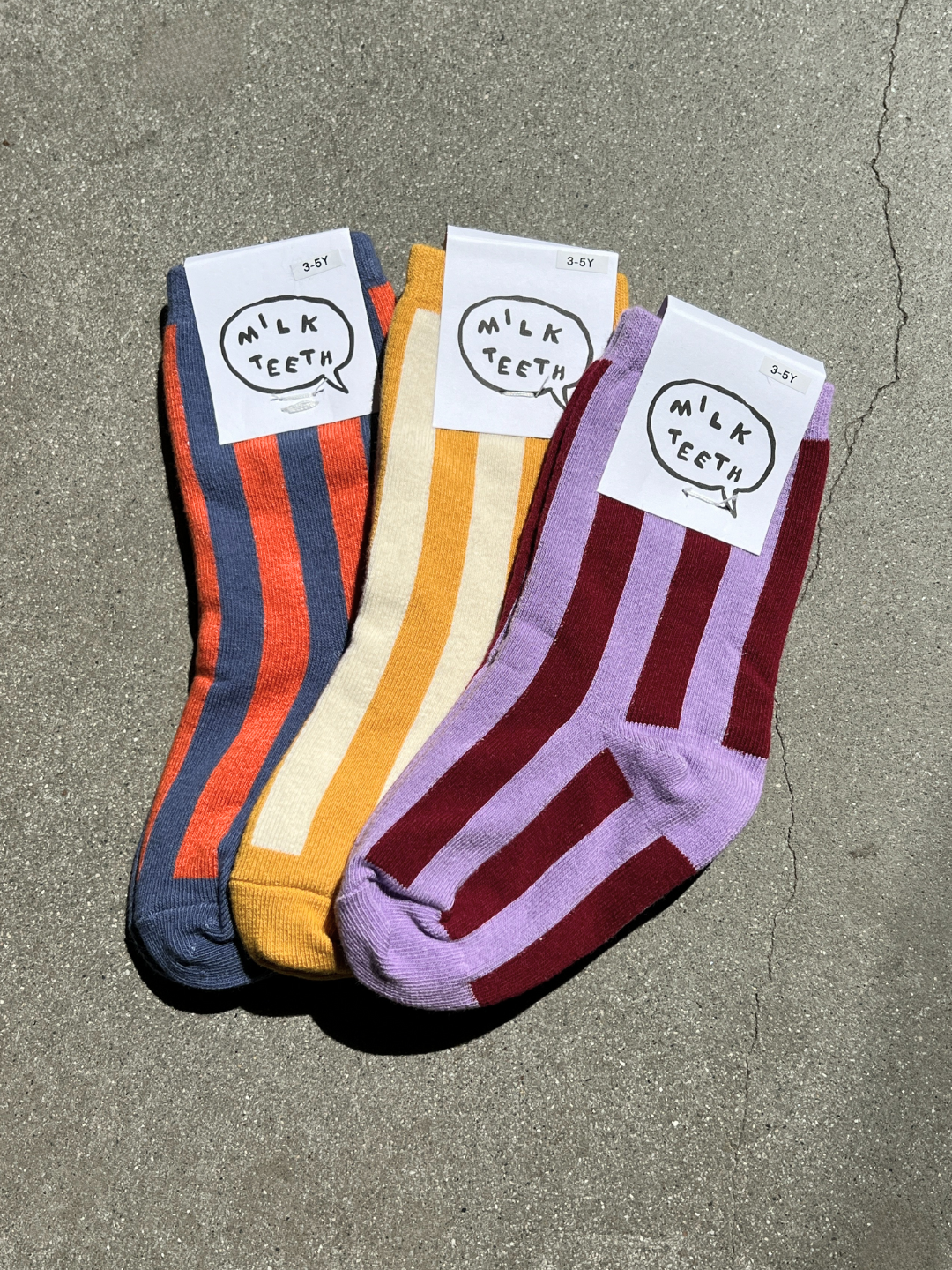 Violet | Three pairs of comfy STRIPE SOCK, featuring bold stripes, are arranged on a concrete surface. Each pair is made from stretchy cotton and displays a tag with the text "Milk Teeth." The colors include blue/orange, yellow/cream, and purple/maroon.