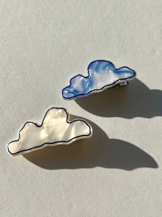 Image of Two CLOUDS HAIRCLIPS, one in pearlescent white and the other in light blue, rest on a light gray surface with shadows cast beneath them. These hairclips feature alligator clip backings for versatility with various hair types.