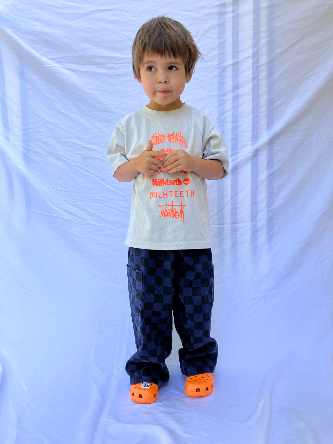 Cement | A child wearing a light grey t-shirt with Milk Teeth spelled out in various fonts in orange ink. He wears navy and black checkered pants and orange Crocs and is standing in front of a white sheet backdrop.