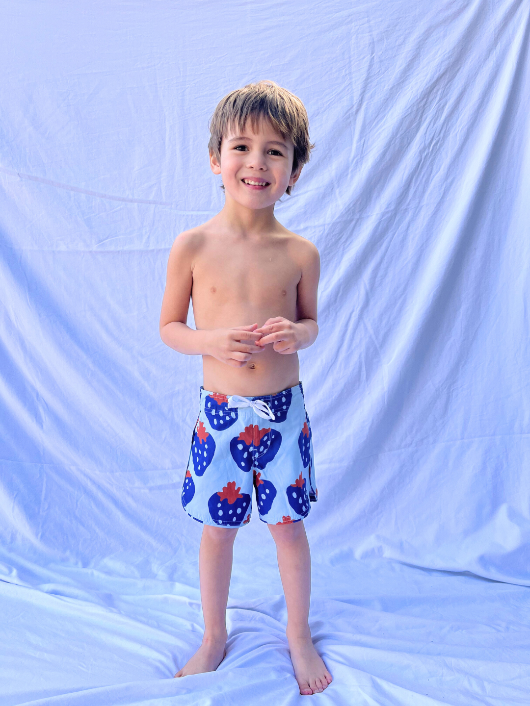 A young boy stands barefoot on a light blue backdrop, wearing STRAWBERRY BOARDSHORTS with red heart patterns. He is smiling, has short brown hair, and radiates pure joy.