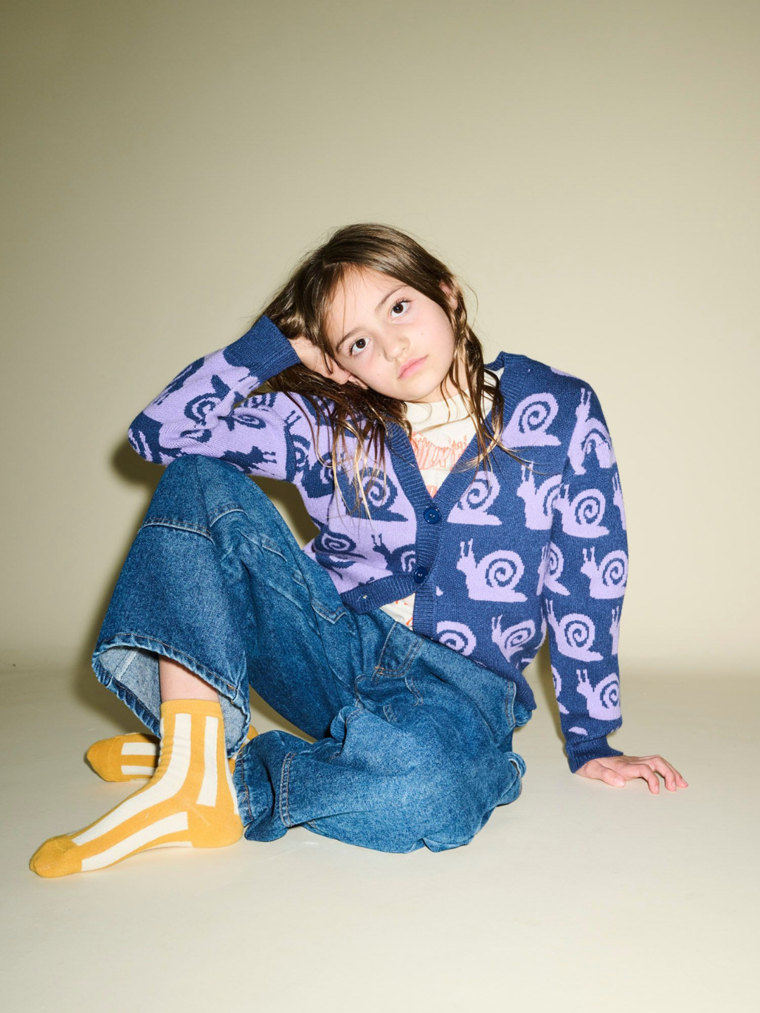 A child sits on the floor against a plain background, wearing an ESCARGOT CARDIGAN, jeans, and yellow striped socks. Gazing intently at the camera, this image is perfect for product pairs showcasing cozy winter wear.