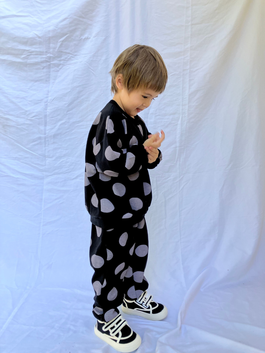 A child wearing a sweat set in black with large grey polka dots. The set consists of a crewneck sweatshirt and a matching sweatpant, with an oversized fit. He wears black velcro sneakers with white trim and is standing on a white sheet backdrop.