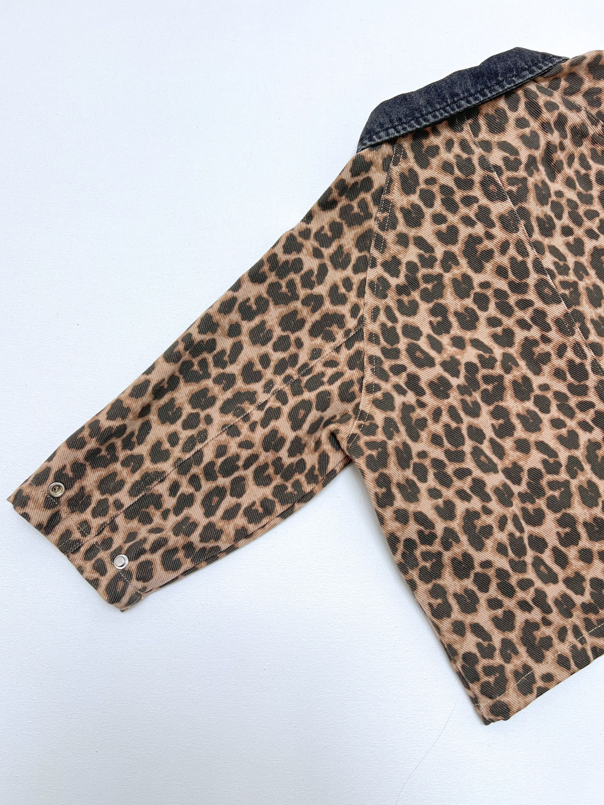 A close-up of a leopard print shirt jacket featuring a tan and brown animal pattern with a dark denim collar and buttoned cuffs, laid flat on a white background.