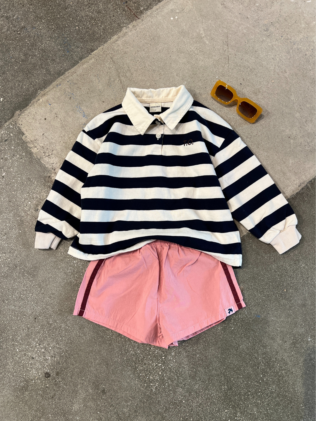 Cream | The RUGBY LONGSLEEVE, pink shorts, and yellow sunglasses from a Korean kids brand are neatly arranged on a concrete surface.