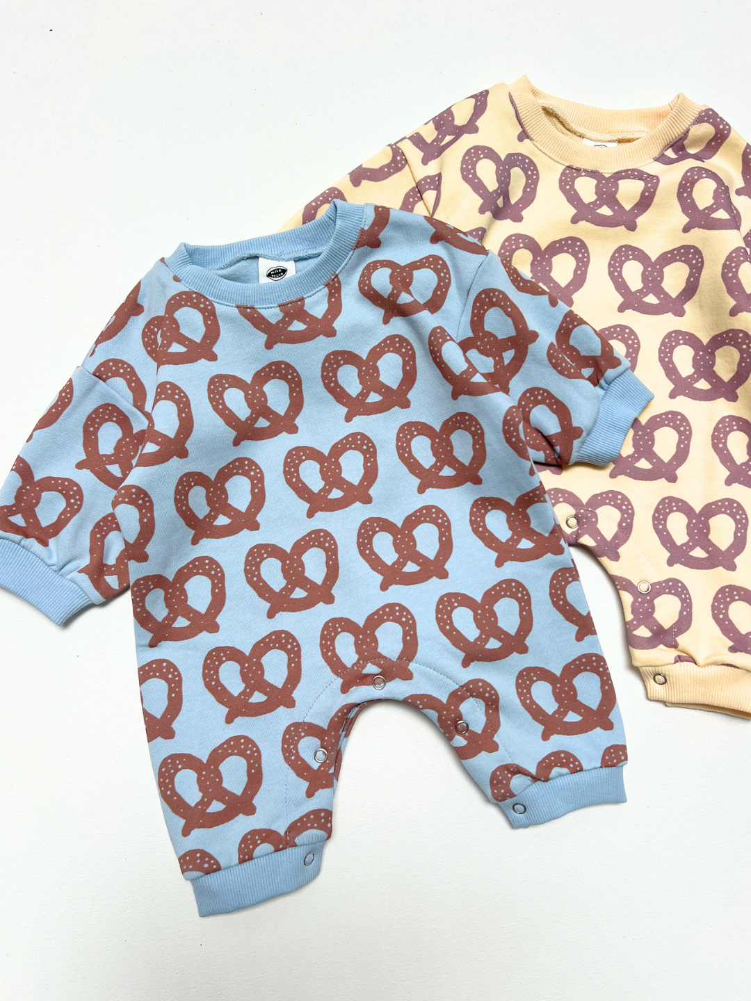 Brown/Blue | Two baby long-sleeve footless onesies laid flat on a white background. The front one is light blue with a repeating brown pretzel print, and the one behind it is pale yellow with a repeating mauve pretzel print.