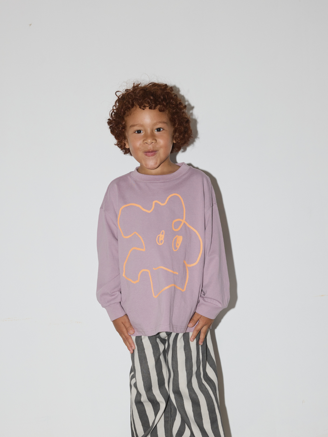 Purple | A young child with curly brown hair wearing a lavender long-sleeve sweatshirt with a yellow cartoon outline graphic on the front, paired with black and white vertically striped pants, standing against a plain light gray wall.