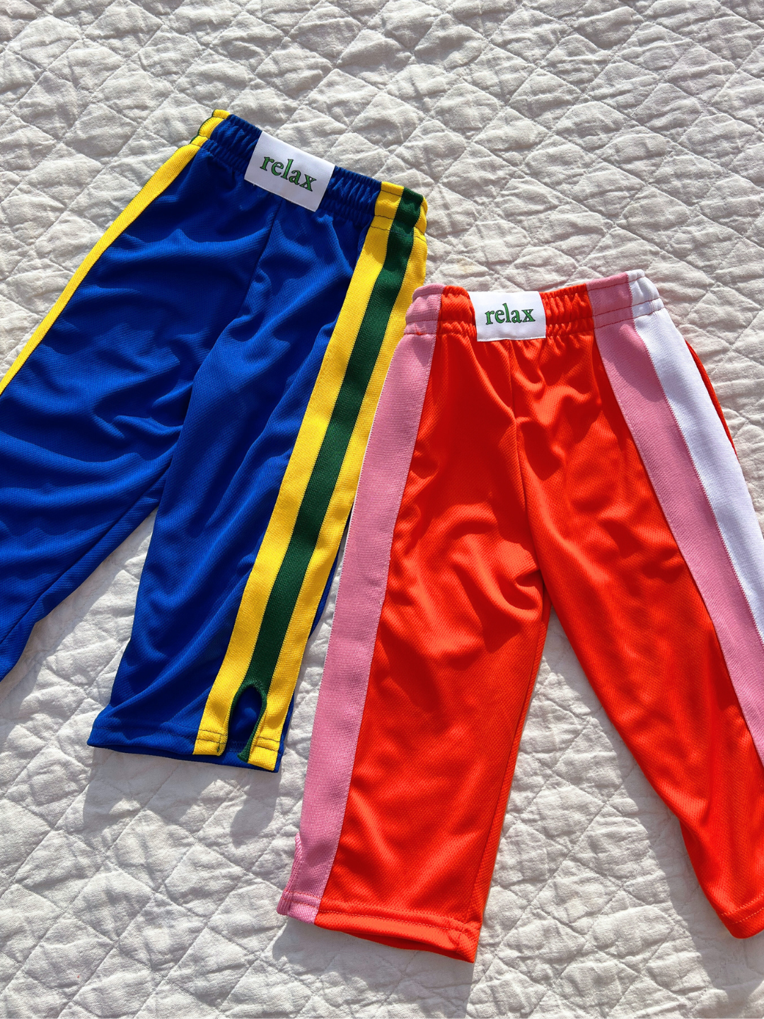 Two pairs of RELAX PANTS for kids rest on a quilted surface: one pair is blue with yellow and green stripes, while the other is red with pink and white stripes. Each pair features "relax" labels on the waistband, crafted from Miracle Mesh fabric for ultimate comfort by Relax Lacrosse Brand.