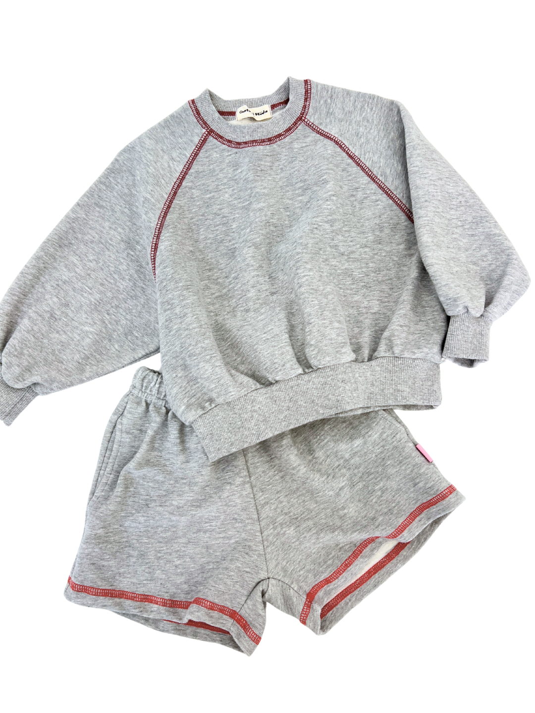 Grey | Cantucci Studios introduces the VARSITY SWEATSHIRT set, a retro-inspired ensemble crafted from soft, brushed cotton. The gray sweatshirt and shorts feature red stitching accents, a crew neck, and an elastic waist for optimal comfort and style.