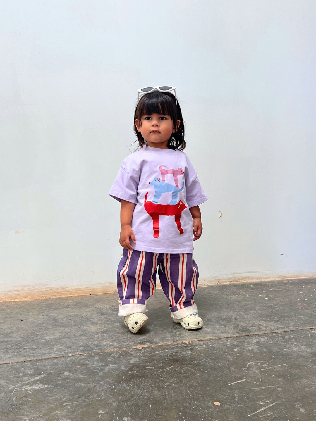 A young child stands indoors wearing a purple animal-print t-shirt, STRIPE TWILL PANT with a baggy fit, white clogs, and sunglasses perched on their head.
