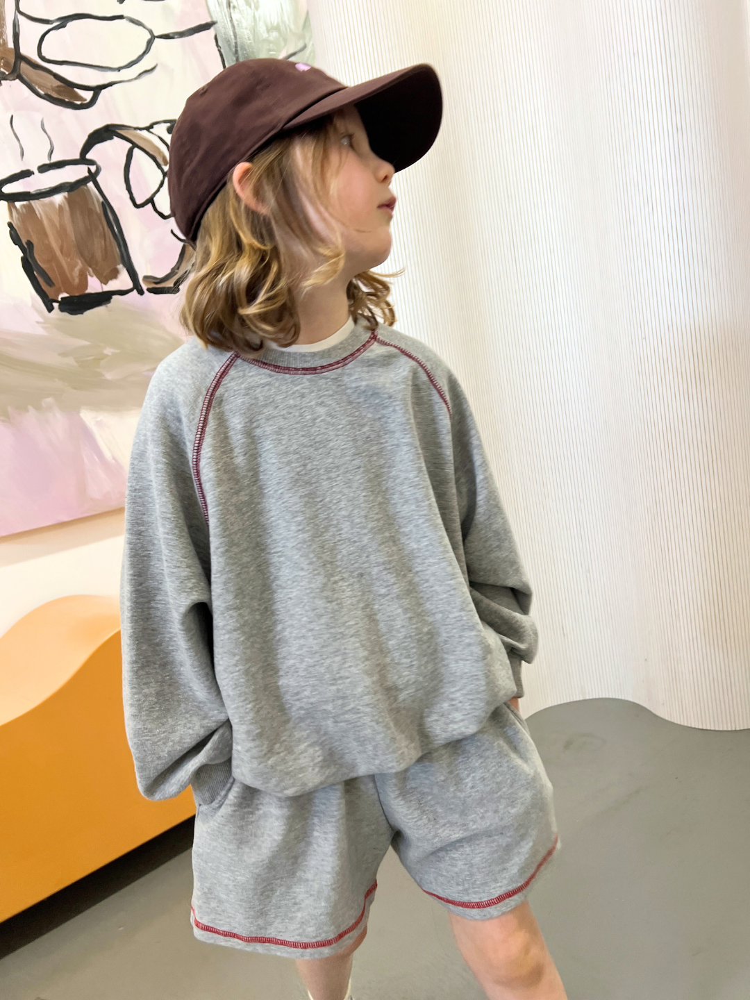 Grey | A child in a brown cap and retro-style gray brushed cotton VARSITY SWEATSHIRT, paired with matching shorts, stands in a room featuring abstract artwork from Cantucci Studios on the wall.