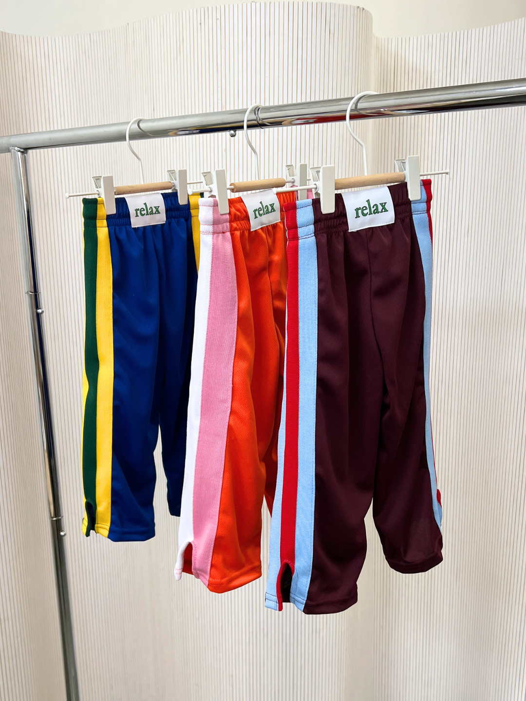 Blue | Four pairs of vibrant RELAX PANTS with side stripes hang on a rack, each adorned with a "relax" label. Displaying bold colors like blue, red, orange, and maroon, these kids' track pants are crafted from Miracle Mesh fabric by Relax Lacrosse Brand to ensure comfort and style for every playful adventure.