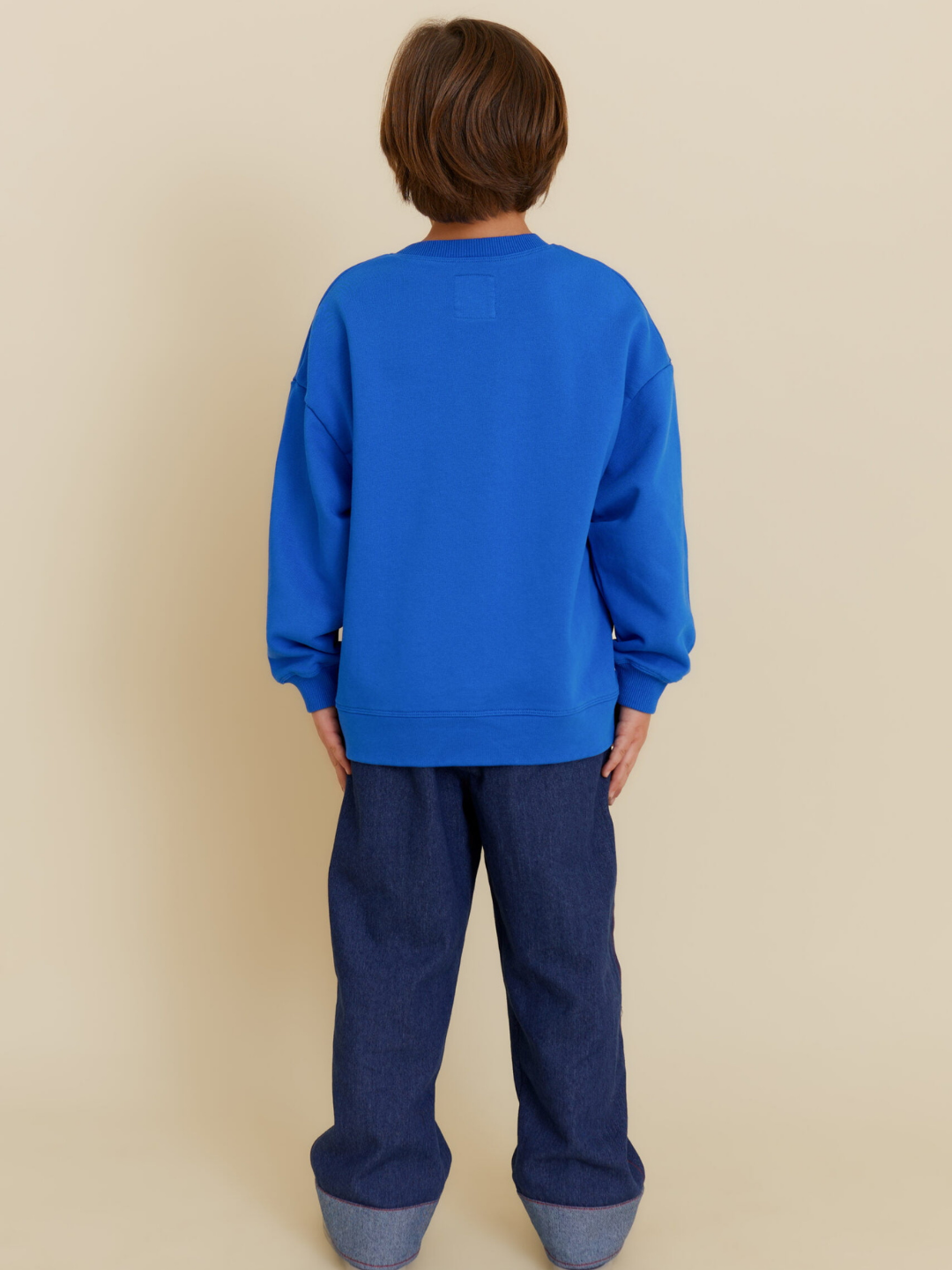 A child faces away from the camera, wearing the OXOX CLUB POTATO SWEATSHIRT in bright blue with dark blue jeans, both made from soft organic cotton, posed against a beige background.
