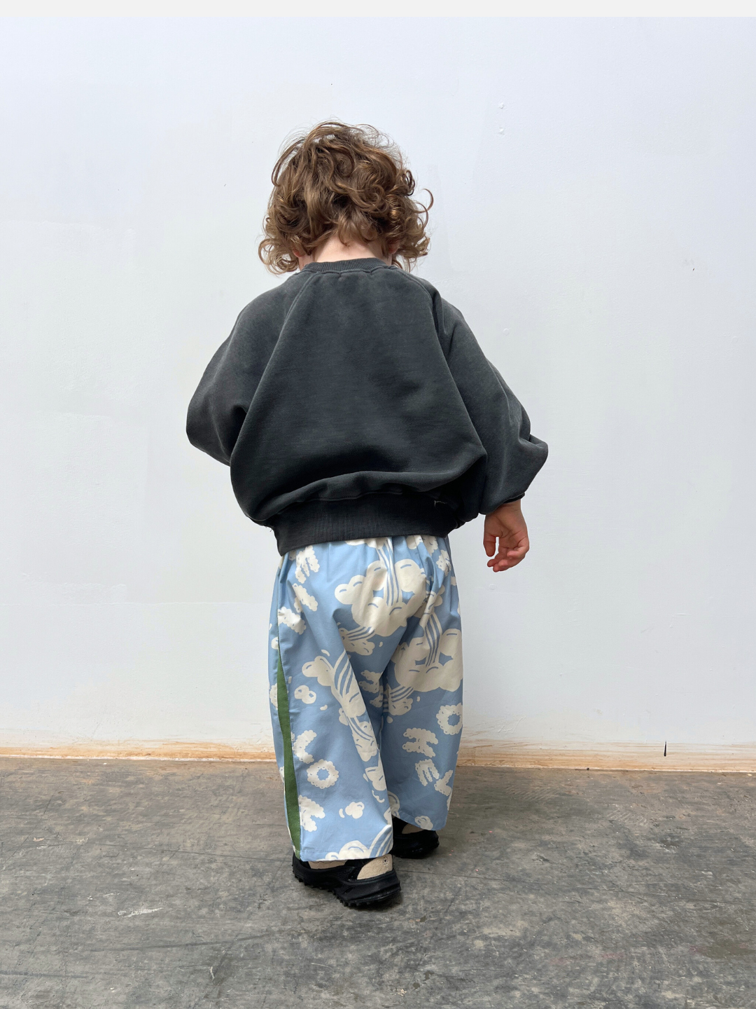 A young child with curly hair stands with their back to the camera, wearing a black sweater and SKYWRITING PANT—loose blue pants featuring a white pattern.
