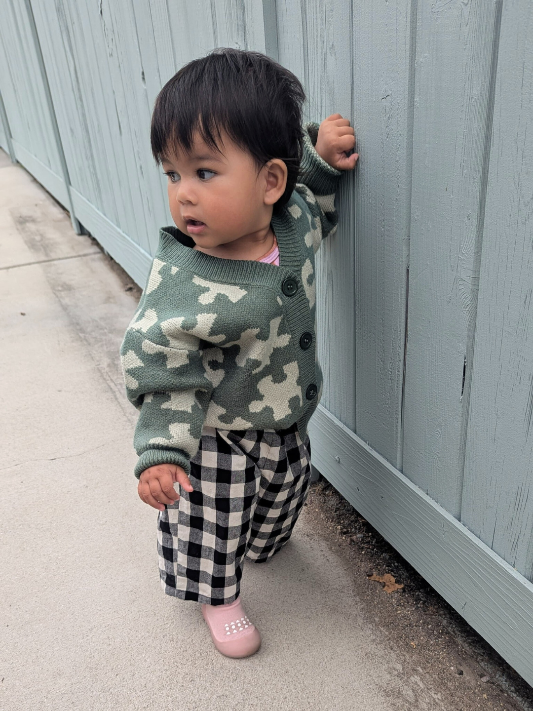 Green | A toddler wearing the PUZZLE CARDIGAN, featuring a green base with white patterns, paired with checkered pants and pink shoes, stands by a light blue wooden fence and looks to the side.