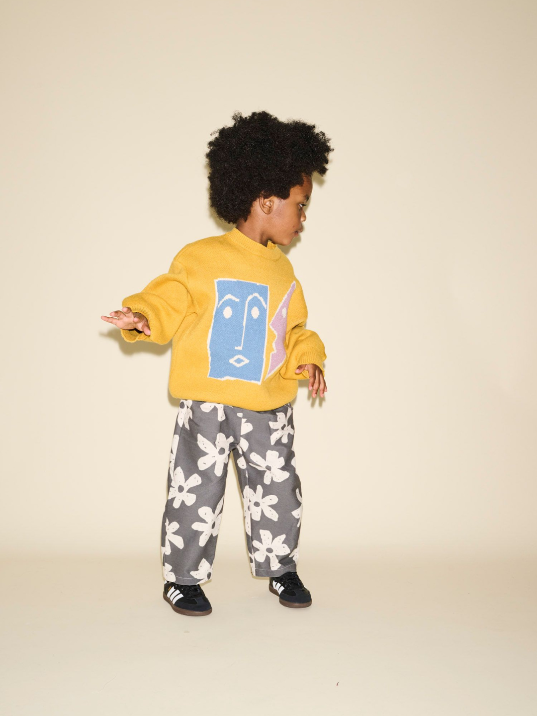 A young child with curly hair wearing the TEATIME SWEATER, featuring a graphic face design on a yellow background, and black floral pants stands against a plain background, perfect for product photos showcasing adorable fashion pairs.