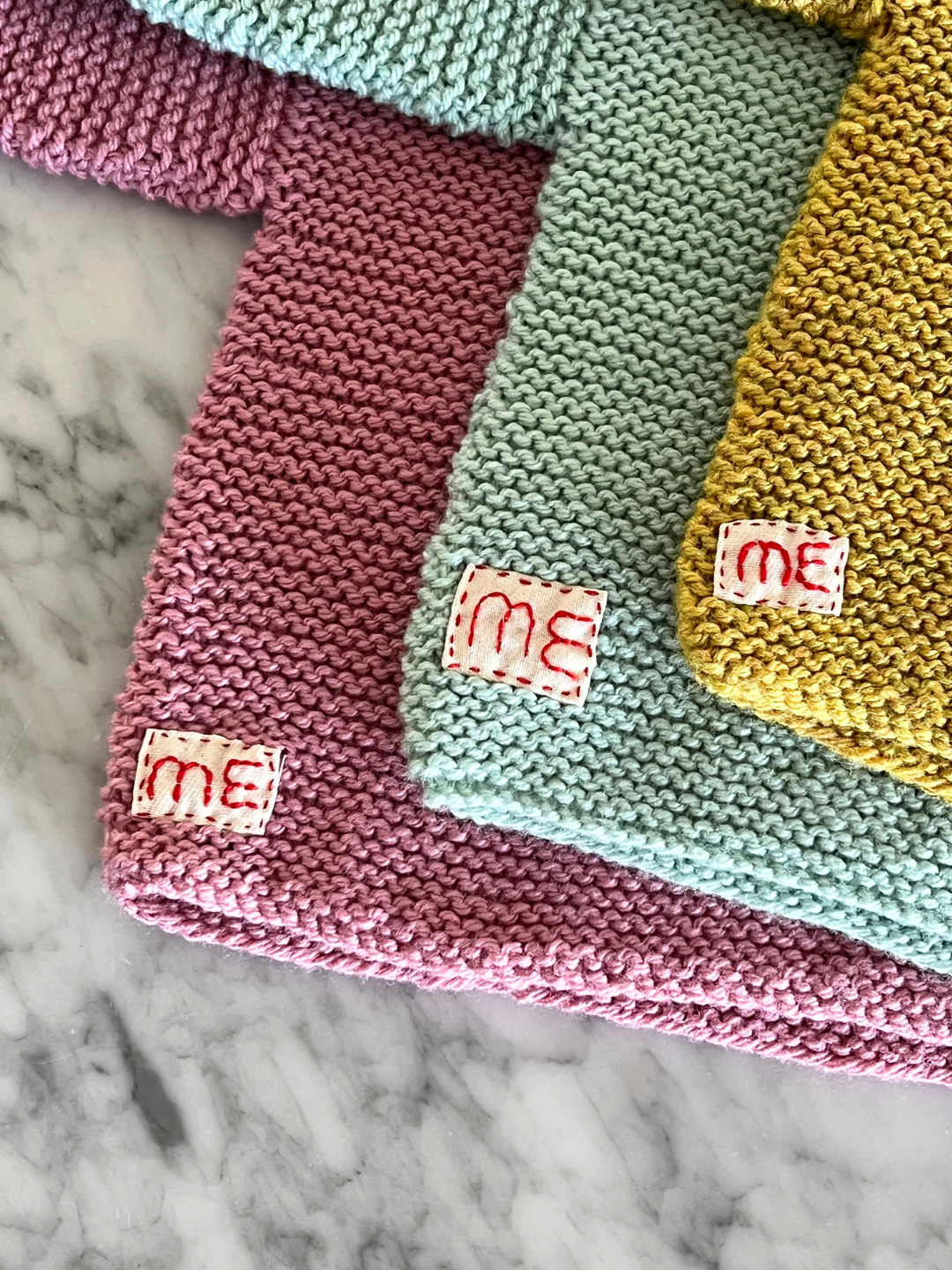 Back detail view of three hand-knitted baby sweaters in pink, mint and ochre, showing the hand-stitched "ME" patch.